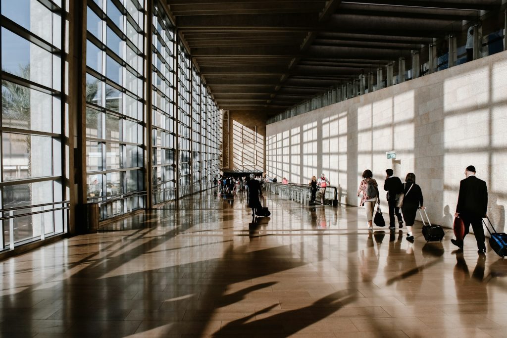 People walking through a sunlit airport terminal with large glass windows and shadows